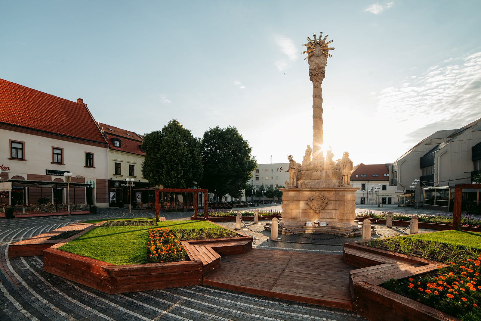 Morový stĺp Najsvätejšej Trojice v Trnave na Trojičnom námestí pri západe slnka, obklopený modernou mestskou zeleňou a kvetinovou výsadbou.