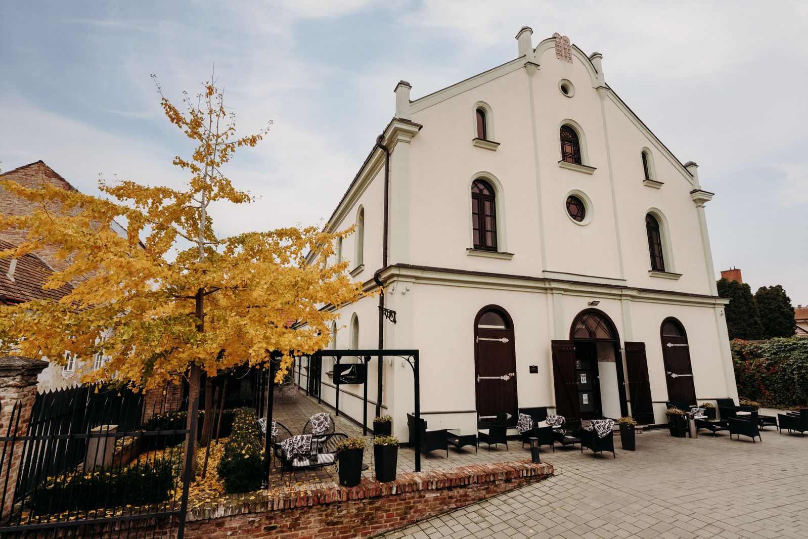 Zrekonštruovaná kaviareň synagóga v Trnave s vonkajším posedením, obklopená jesennou prírodou a žltnúcim stromom, predstavujúca kultúrnu a historickú pamiatku mesta.