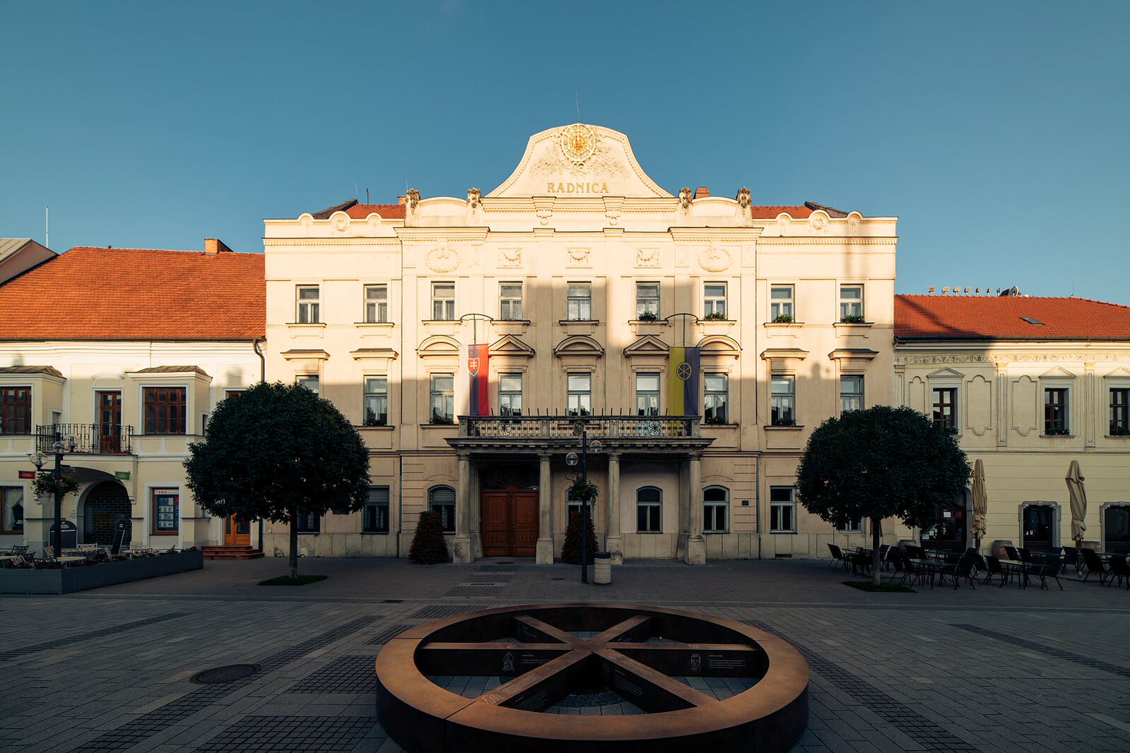 Historická budova radnice v Trnave na Trojičnom námestí s ikonickou sochou mlynského kolesa v popredí, obklopená historickými budovami a zeleňou.