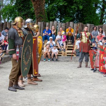 Ukážka rímskych vojakov v archeoparku Cífer-Pác. Niekoľko reenaktorov v historických kostýmoch rímskej armády stojí na pieskovom nádvorí s veľkými štítmi a prilbami, pred skupinou divákov sediacich na drevených laviciach. V pozadí sú drevené palisády a zeleň.