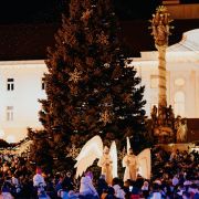 Veľký vianočný stromček na námestí obklopený davom ľudí počas večerného podujatia. Pred stromčekom stoja dvaja účinkujúci v bielych kostýmoch s veľkými anjelskými krídlami. Osvetlené historické budovy tvoria pozadie scény.