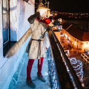 Osoba v historickom kostýme stojí na balkóne vysoko nad mestom a drží v ruke zapálenú lampášovú lampu. Pod ňou sa rozprestiera večerné mesto so svetielkami a vianočnou výzdobou. Scéna pôsobí slávnostne a pripomína tradičný zvyk nočného hliadkovania.