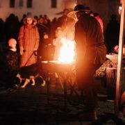 Muž v dobovom oblečení pracuje pri otvorenom ohni na malom kovovom ohnisku. Okolo neho stoja zimne oblečení návštevníci, ktorí sledujú ukážku remesla. Teplé svetlo plameňov osvetľuje scénu uprostred večerného zimného trhoviska.
