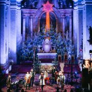Koncert v kostole s bohatou vianočnou výzdobou. Pred oltárom so žiariacou hviezdou vystupuje hudobná skupina s viacerými spevákmi a hudobníkmi. Chrámový priestor je osvetlený modrým a fialovým svetlom, čo vytvára slávnostnú atmosféru.
