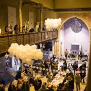 V priestrannej historickej synagóge prebieha komunitné podujatie. Pri dlhých stoloch stoja a sedia ľudia, ktorí si prezerajú výrobky a rozprávajú sa. V priestore visia veľké biele balóny a dekorácie v tvare hviezd. Na galérii aj na prízemí je mnoho návštevníkov, čo vytvára živú, trhovú atmosféru.